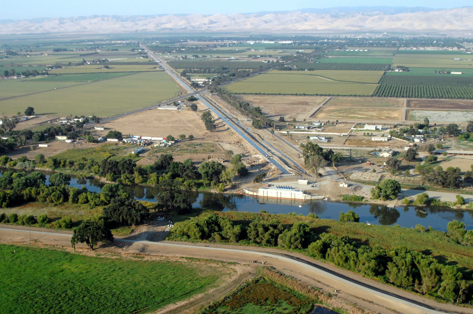 Aerial view of a rural landscape with fields, a river, a small dam, a road, and distant mountains under a clear sky.