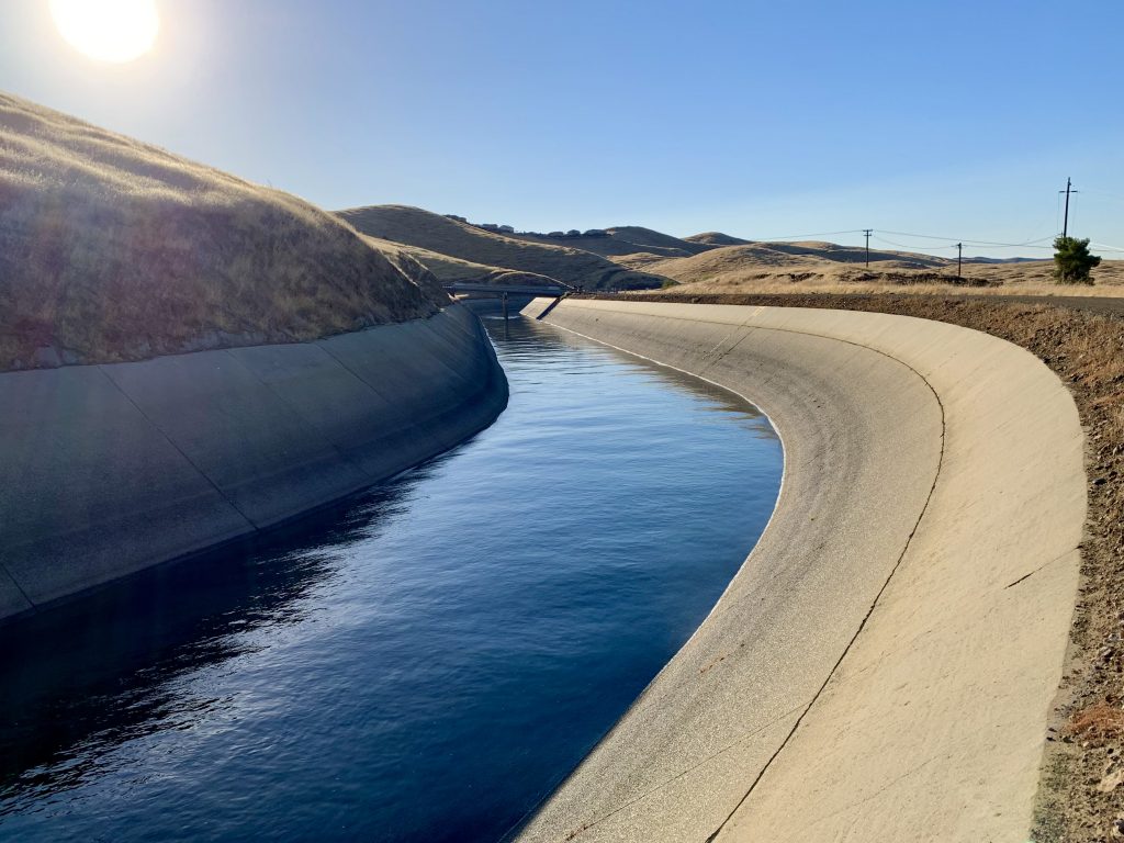 Image of the Friant-Kern Canal, filled with water and headed into a curve with the rising sun behind.