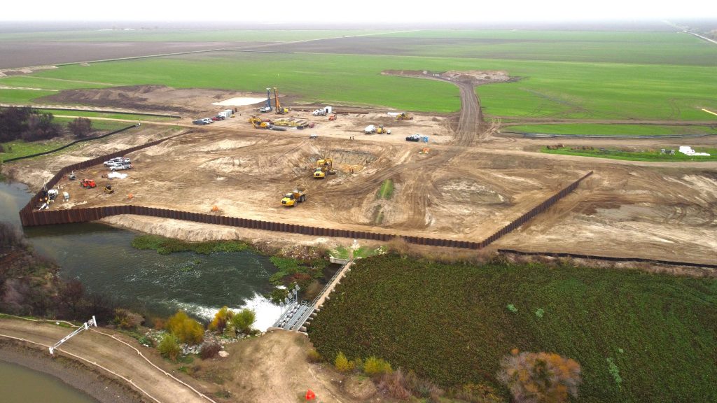 An aerial view of cleared riverbank with heavy machinery and a wall of sheet piling encircling the project site.