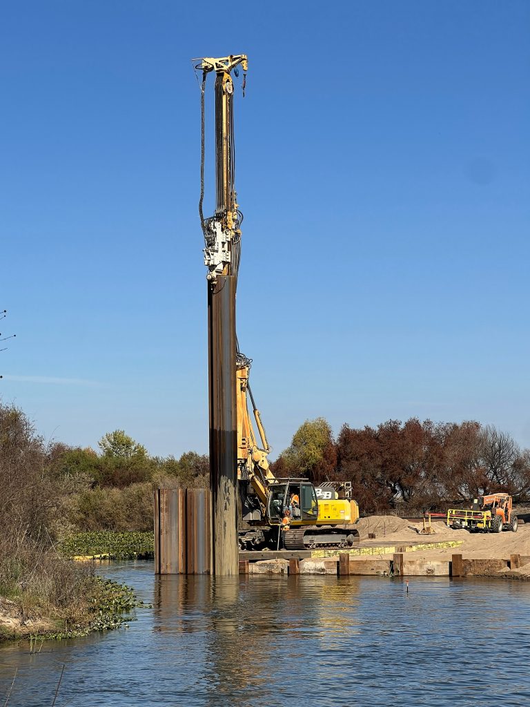 A vibrating hammer drill rig is used to drive sheet piles into the riverbank in order to protect the project site from water.